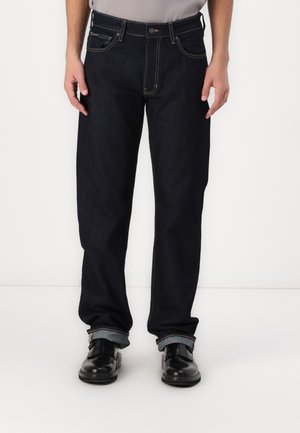 Jeans de mezclilla azul oscuro con corte recto, que presentan costuras en contraste, diseño de cinco bolsillos y dobladillos remangados. Combinados con zapatos negros.