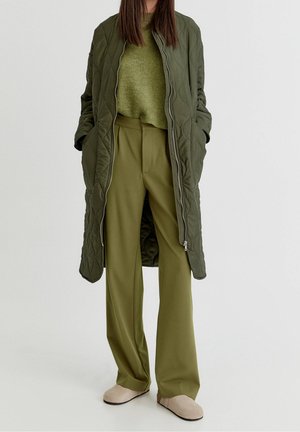 Kobieta ubrana w długi pikowany oliwkowozielony płaszcz nałożony na zielony sweter oraz szerokie oliwkowozielone spodnie, w beżowe buty wsuwane, stoi z rękami w kieszeniach.