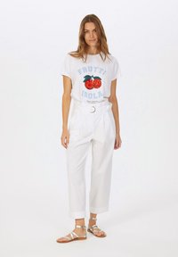 White graphic t-shirt with blue text and red fruit design, paired with high-waisted white trousers featuring a belt and silver sandals.