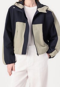 Marinblå och ljusgrå vindtät jacka med dragkedja fram, elastiska ärmslut, dubbel bröstficka och huva. Lättviktig tyg med avslappnad passform.