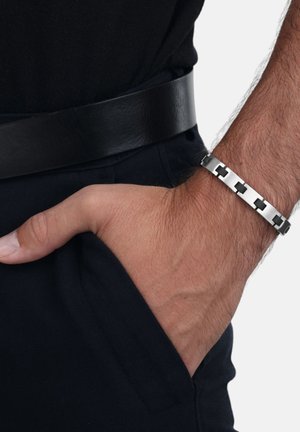 Bracciale in metallo con link geometrici intrecciati, finitura argentata lucida, indossato a un polso, con una cintura in pelle nera visibile sullo sfondo.