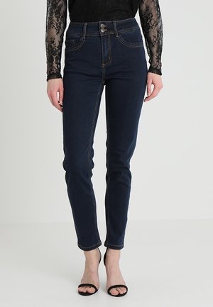 Kvinde iført mørkeblå høj talje skinny jeans kombineret med sort blonder langærmet top og sorte åbne sandaler med hæl.
