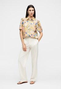 Γυναίκα με μακριά σκούρα μαλλιά που φοράει κοντομάνικο εμπριμέ μπλουζάκι, λευκό παντελόνι με φαρδιά πόδια και σανδάλια, στέκεται με το ένα χέρι στην τσέπη.