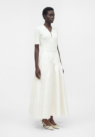 Femme portant un haut blanc à manches courtes côtelé avec col et une longue jupe blanche fluide, associée à des chaussures à petit talon couleur crème.