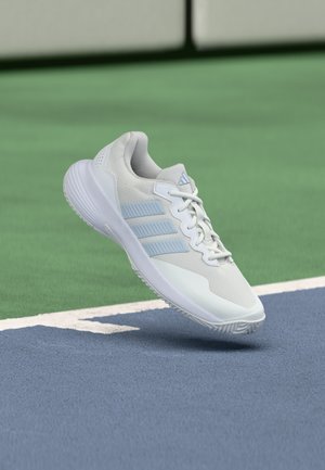 Baskets blanches Adidas avec des rayures bleu clair flottant au-dessus d'une surface de court de tennis bleue et verte près du filet.