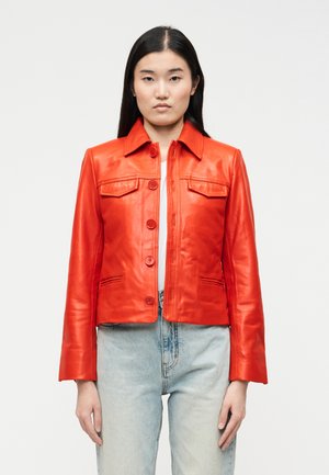 Kobieta w jasnoczerwonej skórzanej kurtce, białej bluzce i jasnoniebieskich dżinsach, stojąca na tle jednolitego białego tła.