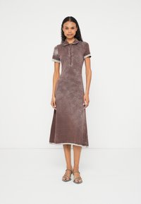 Žena stojaca, oblečená v hnedom zamatovom midi šate s krátkymi rukávmi, zapínanými na gombíky, a v sandáloch na podpätku so remienkami na jednofarebnom bielom pozadí.