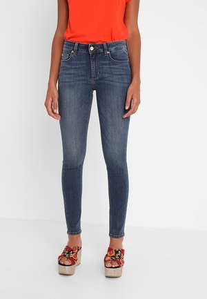 Vrouw draagt donkerblauwe skinny jeans met hoge taille, een oranje top en platformsandalen met bloemenpatroon, staand tegen een witte achtergrond.