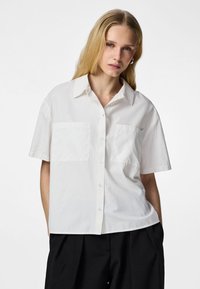 Lyshåret kvinde iført en løs, hvid kortærmet skjorte med knapper og to brystlommer samt sorte vide bukser mod en hvid baggrund.