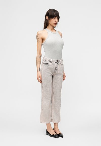 Γυναίκα με τατουάζ που φοράει γκρι αμάνικο τοπ, ξεβάμμένο τζιν με ευρύ πόδι και μαύρες μυτερές μπαλαρίνες, που στέκεται σε ουδέτερη στούντιο ατμόσφαιρα.