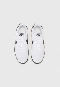 Hvide Nike-sneakers med sorte detaljer, flade snørebånd, polstret tunge og rund tådesign. Fremstillet af kanvas og gummisål.