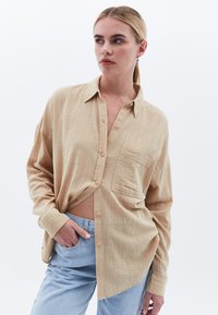 Camicia button-up beige in tessuto texturizzato, con taschino sul petto e maniche lunghe, abbinata a jeans azzurri chiari.