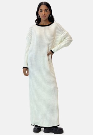 Langes weißes geripptes Strickkleid mit schwarzem Besatz am Ausschnitt und an den Ärmeln, das einen lockeren Schnitt und lange Ärmel hat.