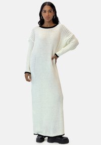 Lang, ribbet hvid kjole med sort kant ved halsen og manchetterne, der har en bred, afslappet pasform og lange ærmer, kombineret med sorte platåstøvler.