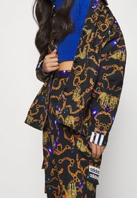 Svart mönstrad jacka och byxset med guldkedjor och blommotiv, kompletterat med en blå stickad tröja under.