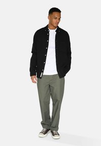 Sort skjorte med knapper over en hvid T-shirt, kombineret med olivenfarvede bukser og brune sneakers. Uden syninger lommer på skjorten og bukserne.