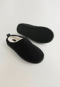 Musta tossu, joka on valmistettu tekstuurisesta, pörröisestä materiaalista, pehmeällä sisävuorella, jossa on kumipohja, jossa on aaltoileva kuvio ja vetolappu.