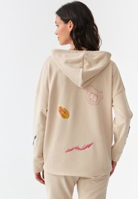 Sudadera beige con capucha y diseños bordados, que incluyen la cara de un oso y motivos coloridos, con un ajuste relajado y una textura suave.