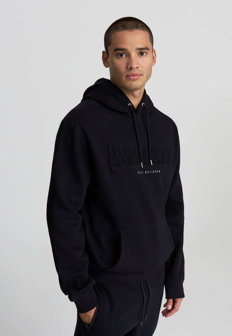 Giovane uomo che indossa un felpa nera con cappuccio e pantaloni con la scritta "BREAKER SELF BELIEVER", in piedi su uno sfondo grigio chiaro uniforme.
