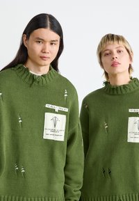 Pulls en tricot vert avec des ourlets effilochés, des déchirures proéminentes et une poche avec des détails de marque. Tissu texturé, design surdimensionné.