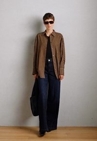 Chemise oversize à carreaux marron, haut noir, jean flare en denim foncé taille haute, sac à main noir, lunettes de soleil foncées, posant contre un fond gris.