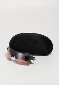 Sportliche Sonnenbrille mit einem dunkelgrauen Rahmen und rosa getönten Gläsern, begleitet von einem schwarzen Hartschalenetui mit Logo.