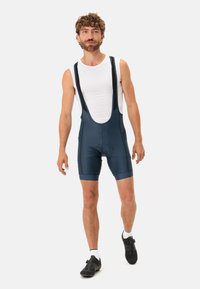 Jūras spēku velosipēdu bikses ar regulējamiem siksnām, ar mīkstinātu sēžamvietas daļu un gludu, elastīgu audumu. Valkātas virs baltas piegulošas krekla.