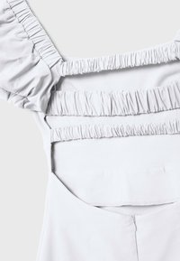 Vêtement gris clair avec un volant élastique le long de l'encolure et dans le dos. Texture de tissu lisse avec une coupe ajustée et des détails de couture subtils.