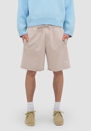 Osoba ubrana w beżowe szorty ze sznurkiem, niebieski sweter, białe skarpetki oraz beżowe sznurowane buty, stojąca z rękami w kieszeniach.