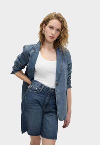 Blazer in denim di un blu medio con maniche lunghe, abbinato a pantaloni corti in denim a vita alta dello stesso tono e orlo grezzo. Top bianco sotto.