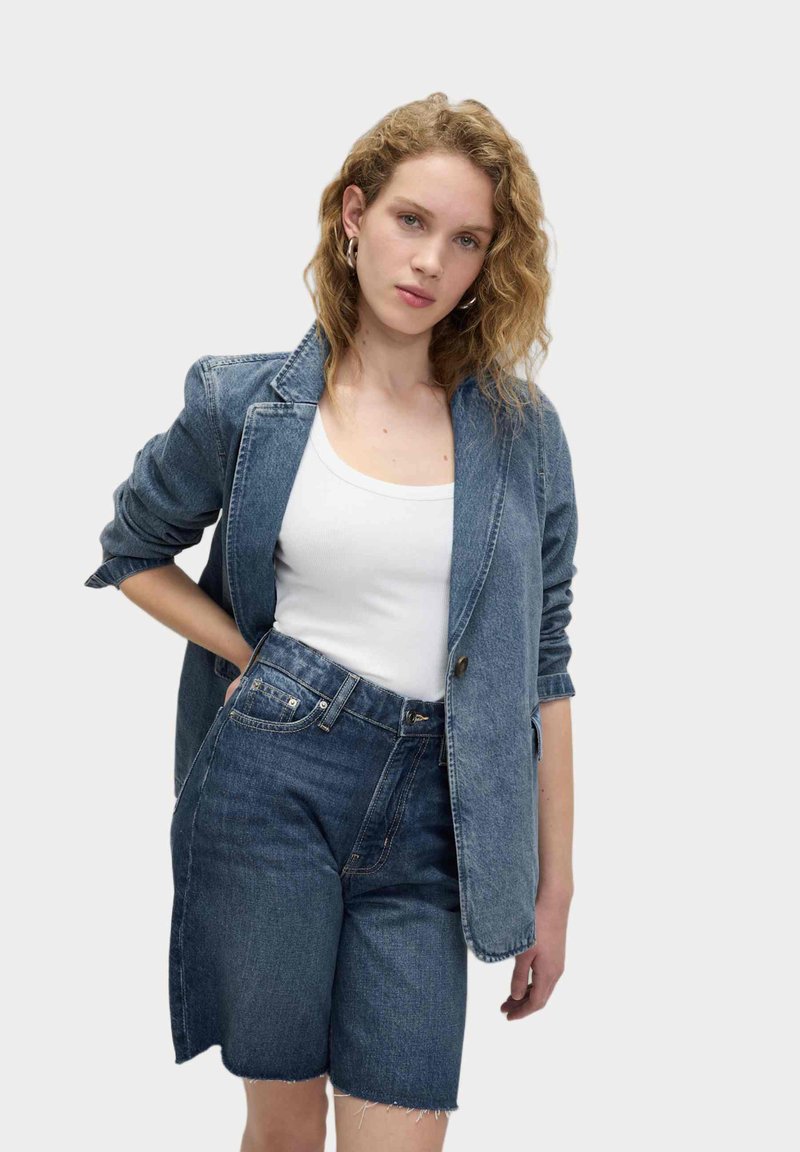 Blazer in denim di un blu medio con maniche lunghe, abbinato a pantaloni corti in denim a vita alta dello stesso tono e orlo grezzo. Top bianco sotto.