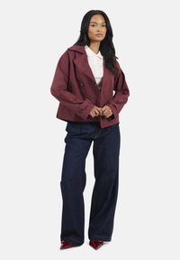 Manteau en trench coupé, bordeaux, avec de larges revers et un design à double boutonnage, associé à un jean bleu foncé ample et des escarpins rouges à bout pointu.