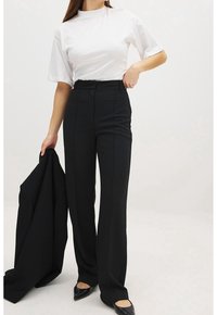 Schwarze maßgeschneiderte Hose mit hoher Taille und geradem Bein, kombiniert mit einem weißen Kurzarmtop. Flache schwarze Schuhe und ein Blazer werden gehalten.