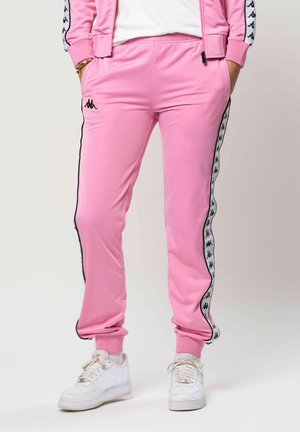 Isik, kes kannab roosaid dressipükse musta-valge logoribaga külgedel ja valgeid vabaajajalatseid, seisab käed taskutes.