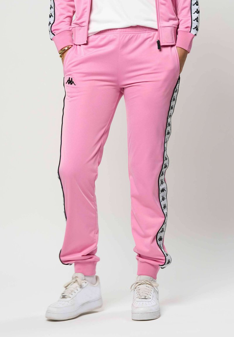 Osoba v ružových teplákoch s čierno-bielym logom po bokoch a bielych teniskách, stojaca s rukami v vreckách.