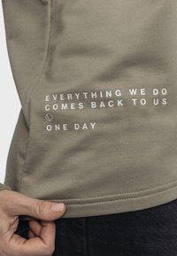 Olivegrüner Sweatshirt mit glatter Textur, das weißen Drucktext auf dem Saum trägt: „Alles, was wir tun, kommt eines Tages zu uns zurück.“