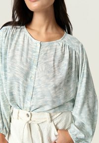 Blusa azzurra chiara con un motivo testurizzato, scollo rotondo, chiusura con bottoni e maniche leggermente arricciate; abbinata a pantaloni bianchi a vita alta con texture.