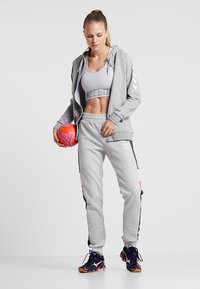 Un ensemble de sport gris comprenant un sweat à capuche zippé et un jogging, avec un soutien-gorge de sport gris court arborant un logo. Tenant un ballon de soccer rose.