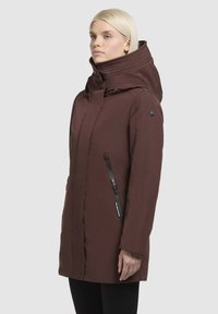 Brūns, garš mētelis ar augstu apkakli un kapuci, ar rāvējslēdzēja kabatu un melniem akcentiem. Gluda tekstūra un pieguļošs dizains.