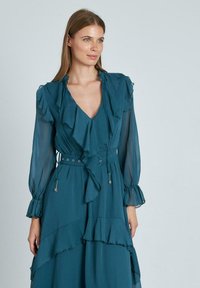 Tealfarbenes Kleid mit geschichteten Rüschen, langen Ärmeln und einem V-Ausschnitt. Verfügt über eine taillierte Taille mit einem Gürtel und dekorativen Beschlägen. Leichtes Material.