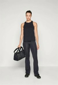 Črn športni top in prilegajoče se hlače iz gladkega, raztegljivega materiala. Model drži črno športno torbo. Preprost dizajn brez vzorcev.