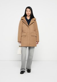 Lichtbruin parka-jack met een capuchon, twee voorzakken en een knoopsluiting. Combinatie met wijde grijze broek en een witte handtas.