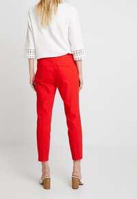 Pantalon rouge sur mesure avec une coupe élégante, associé à un haut blanc avec des découpes sur les manches, et des chaussures à talons blocs beiges.