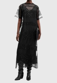 Robe noire transparente avec des rayures pailletées, manches courtes, et texture superposée, associée à des bottes noires jusqu'au genou et à une pochette.