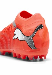 Kirkkaan oranssi jalkapallokengä, jossa on kuvioitu päällinen, mustavalkoinen PUMA-logo ja punaiset nappulat. Kevyt malli, joka tarjoaa pitoa ruoholla.