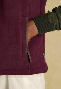 Fleece bunda v tmavej burgundskej farbe, s vrecúškom na zips s hnedým lemovaním, rýhami na tmavozelených manžetách a mäkkou textúrou.