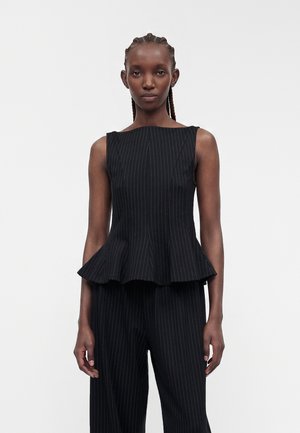 Femme aux cheveux tressés portant un top noir à fines rayures sans manches avec basque et pantalon assorti sur fond blanc uni.