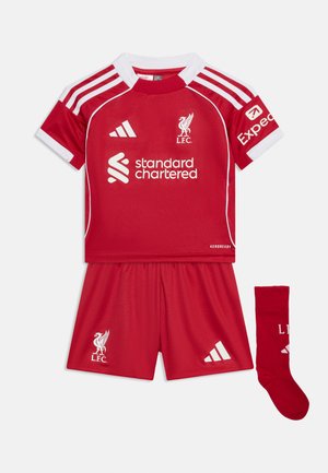 Κόκκινη ποδοσφαιρική εμφάνιση βραχείας μανίκας της Liverpool FC με λευκή επένδυση, που φέρει τα λογότυπα της Adidas και της Standard Chartered, καθώς και κόκκινες κάλτσες που ταιριάζουν με τα αρχικά του συλλόγου.