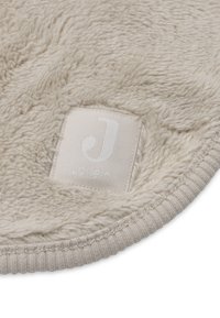 Close-up van beige zachte fleece stof met ribbelrand en een genaaid rechthoekig label met gestileerde "J"-logo en merknaam.