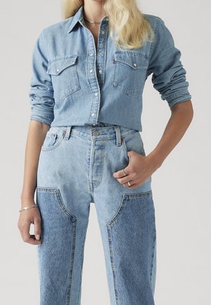 Femme portant une chemise en denim bleu clair avec manches retroussées et un jean denim bicolore, main dans la poche et bijoux en argent.